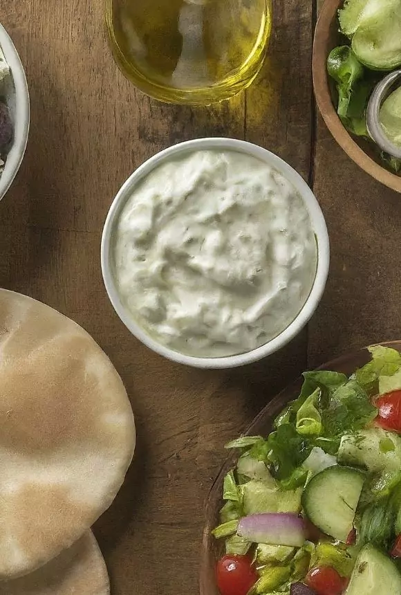 A vibrant Greek bowl featuring creamy tzatziki sauce, fresh vegetables, and grilled meat, elegantly arranged on a wooden table.