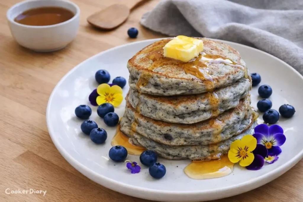 Top-down view of a stack of blue corn pancakes with butter and maple syrup, garnished with fresh blueberries and edible flowers on a wooden countertop.