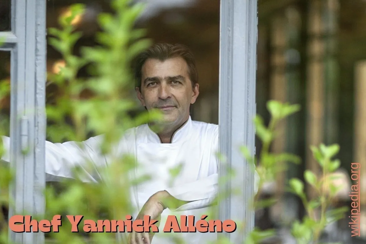 Chef Yannick Alléno standing by a window surrounded by greenery, wearing a white chef's coat.