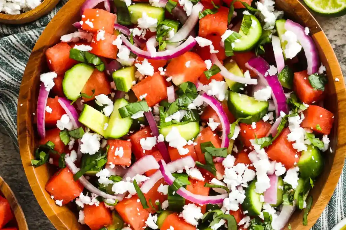 A bright and colorful salad bowl filled with cubed watermelon, sliced cucumber, crumbled feta cheese, and fresh mint leaves, lightly drizzled with a citrus dressing and served on a white ceramic plate with a wooden spoon nearby.
